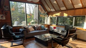 Una sala de estar acogedora con un sofá, dos sillones, una mesita de centro y una chimenea. Amplios ventanales ofrecen vistas al exterior.