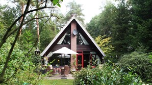 Edificio de madera triangular con puerta roja, rodeado de abundante vegetación y un patio con mobiliario exterior.