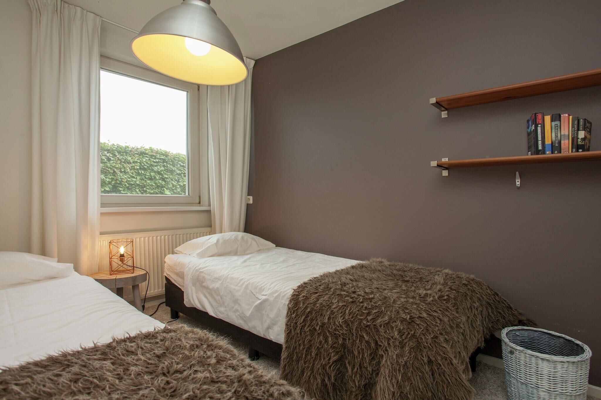 A bedroom with two beds, a window with curtains, a bedside lamp, a radiator, a bookshelf, and a wicker basket.