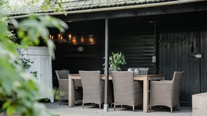 An outdoor dining area with wicker furniture and a table, set against a dark wooden backdrop. - Cottage in Leenderbos with BBQ & Garden (Leende)