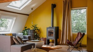 A living room with a sloped ceiling, a wood-burning stove, a sofa, a chair, and a large window. - Cottage in Leenderbos with BBQ & Garden (Leende)