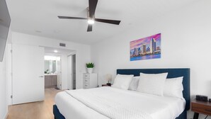 A modern hotel room with a large bed, a ceiling fan, a cityscape painting, and a view of the bathroom.
