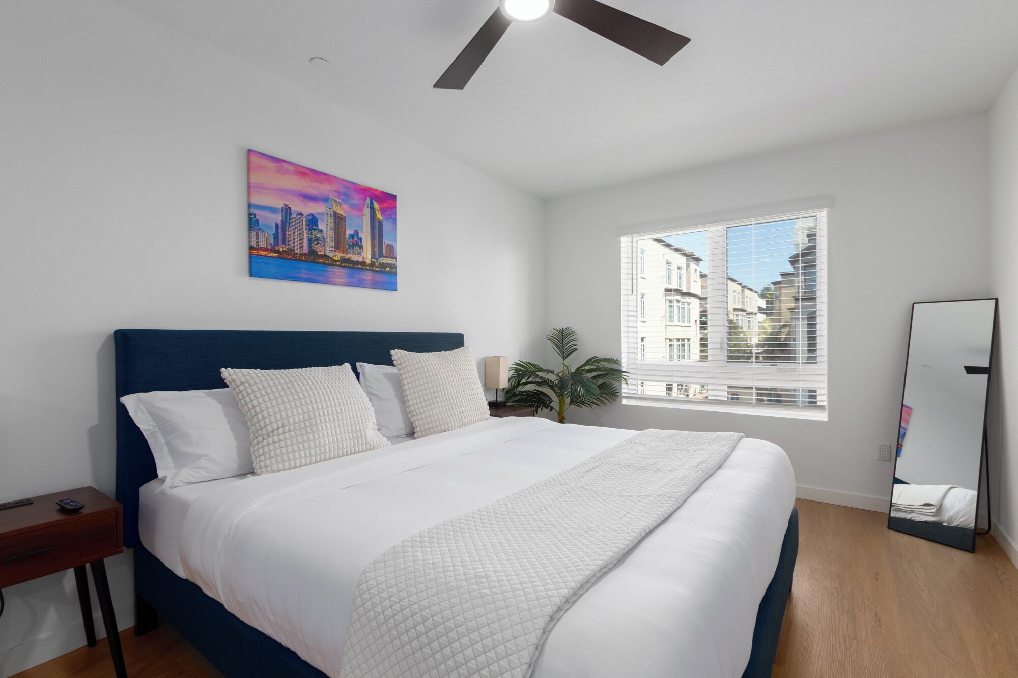 Ein Schlafzimmer mit einem großen Bett, einem Deckenventilator, einem Stadtbildgemälde, einem Fenster mit Blick auf Gebäude und einem Spiegel.