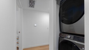 A laundry room with stacked washers and dryers, a vent on the ceiling, and a door leading to a hallway with a shelf of folded towels.