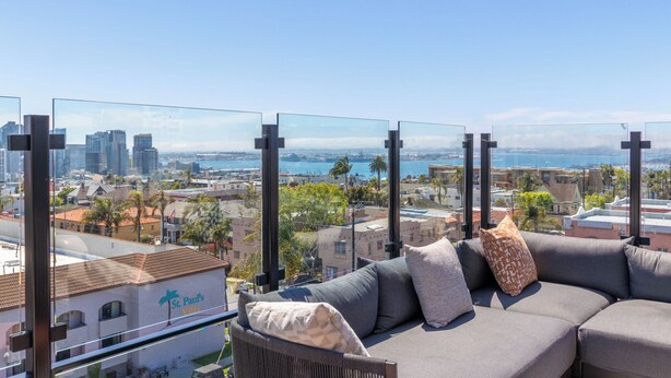 A balcony with a sofa and cushions, overlooking a cityscape with buildings and a body of water.