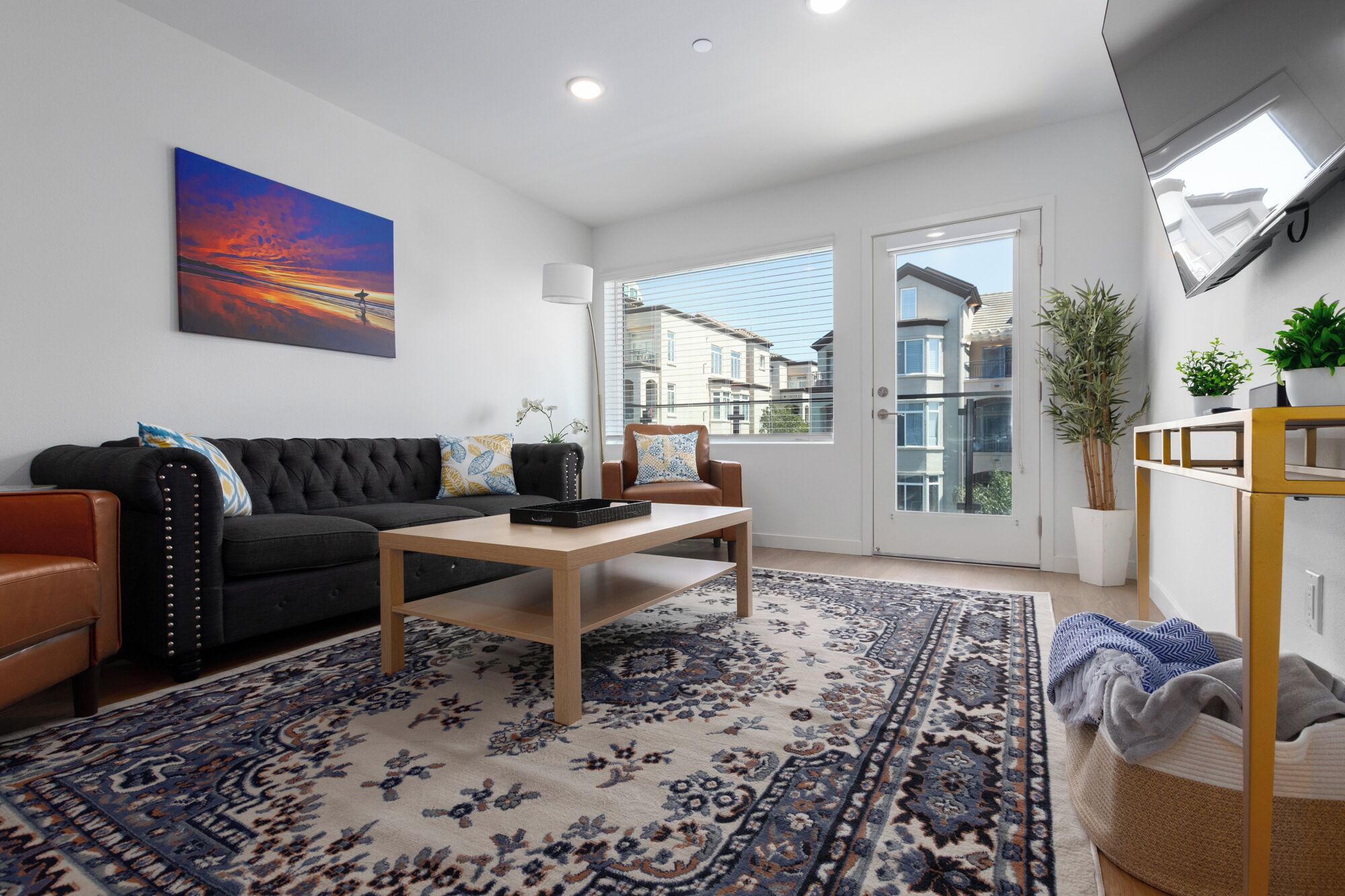 A modern living room with a black sofa, a wooden coffee table, and a patterned rug. There is a large window with a view of buildings outside.