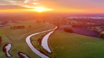 An aerial view of a rural landscape during sunset with fields, a winding water channel, and distant buildings.