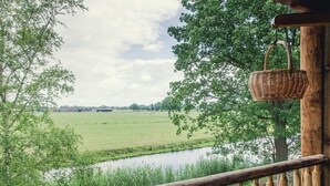 A wooden deck with a wicker basket hanging on a post, overlooking a grassy field and a body of water. - Tranquil Dutch Countryside (Zuna)