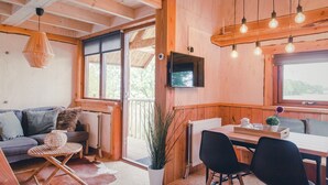 A cozy cabin interior with a wooden ceiling, a dining area with a table and chairs, a sofa with cushions, and a large window providing a view outside. - Tranquil Dutch Countryside (Zuna)