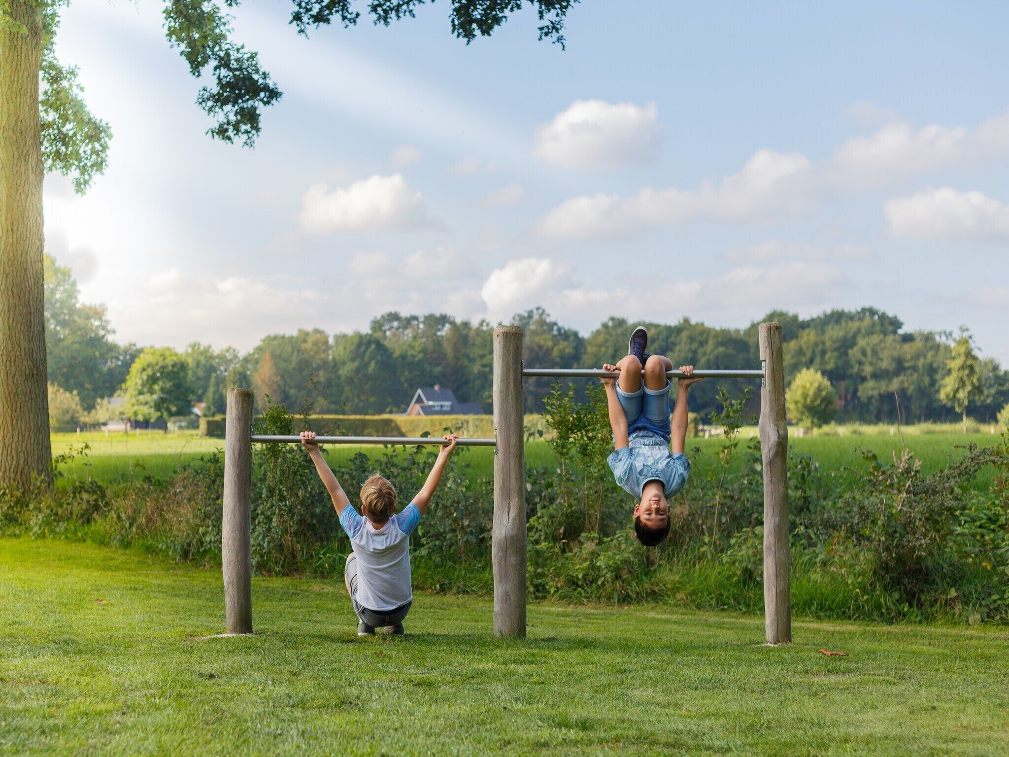 Two individuals performing pull-ups on a horizontal bar in an outdoor setting with a grassy field and trees in the background.