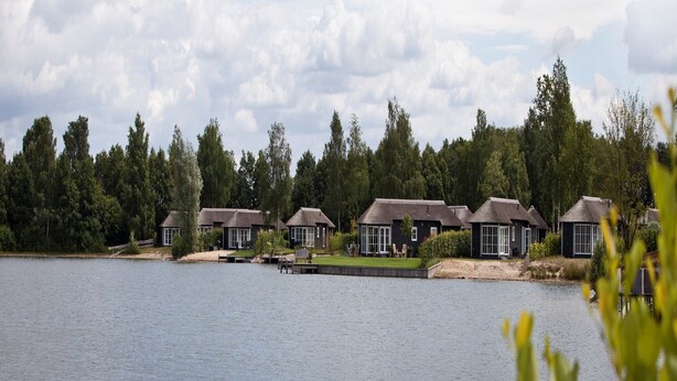 A lakeside hotel with multiple buildings, each featuring a thatched roof and large windows. - Holiday home in park in Twente (Hoge Hexel)