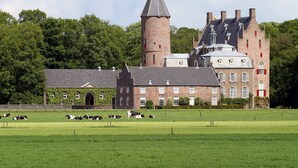 Un edificio grande e histórico con una torre destacada y varias chimeneas, rodeado de un campo verde cuidado donde pastan vacas.