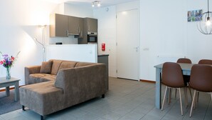 Moderni olohuone, jossa on sohva, ruokapöytä tuoleineen, keittiöalue kaappeineen ja kodinkoneineen sekä eteinen, jossa on ovi.