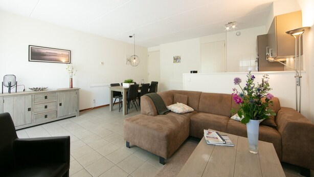 A modern living room with a brown sofa, a dining table, and a kitchen area in the background.