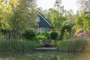 A house with a pond, surrounded by lush greenery and two wooden chairs on a platform. - Charming Home w/ Garden (Paasloo)