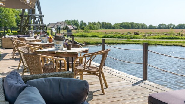 A wooden deck with wicker furniture and a table set for dining, overlooking a calm body of water and a green landscape. - Wellness Villa by the Weerribben (Paasloo)