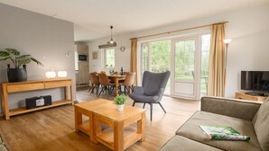 A modern living room with a wooden coffee table, a grey armchair, and a beige sofa. There is a dining area with a table and chairs, and a TV on a wooden stand. - Wellness Villa by the Weerribben (Paasloo)