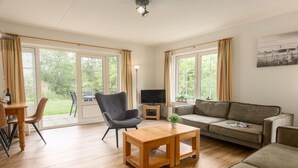 A spacious living room with a wooden coffee table, a grey armchair, a beige sofa, and a television on a stand. Large windows with curtains provide natural light and views of the outdoors. - Wellness Villa by the Weerribben (Paasloo)