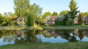 A tranquil lakeside setting with three houses, each with a distinct architectural style, surrounded by lush greenery and a calm pond reflecting the structures. - Wellness Villa by the Weerribben (Paasloo)