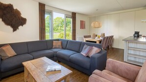 A modern living room with a grey sectional sofa, a wooden coffee table, and a pink armchair. There is a dining area with a table and chairs, and a kitchen area with a counter and hanging light fixture. - Luxury villa with shared pool (Paasloo)