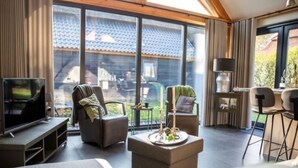 A modern living room with a TV, comfortable seating, a small table with candles, and large windows offering a view of a house with a tiled roof.