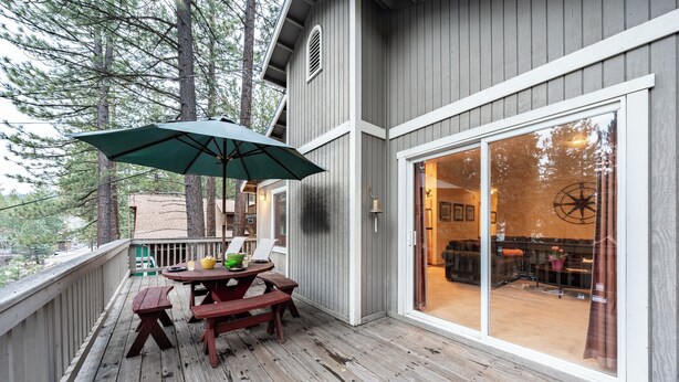 Une terrasse en bois avec des tables de pique-nique et un parasol, une porte-fenêtre coulissante donnant sur un espace intérieur, et une vue sur les arbres en arrière-plan.