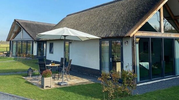 Een modern huis met rieten dak, grote ramen en een terras met tuinmeubilair.
