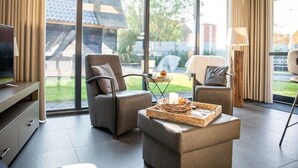 Een moderne woonkamer met een grote schuifdeur die uitkomt op een tuin, een flatscreen-tv, twee leren fauteuils, een salontafel met een dienblad en een lamp.