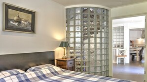 Ein Schlafzimmer mit Bett, Nachttisch, Lampe, einem gerahmten Kunstwerk und Blick auf eine Küche durch eine Glasabtrennung.
