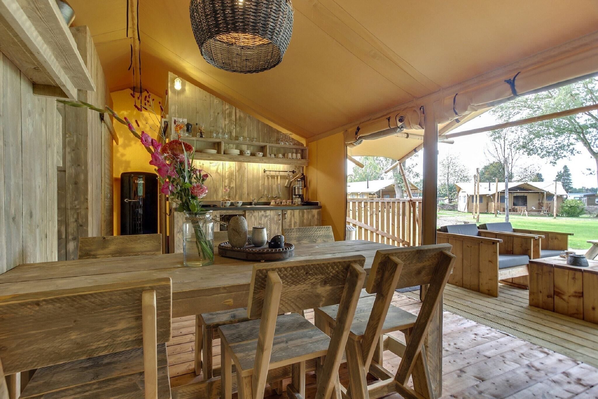 Una terraza de madera con una mesa y sillas de comedor, una zona de cocina con armarios de madera, y una lámpara colgante.