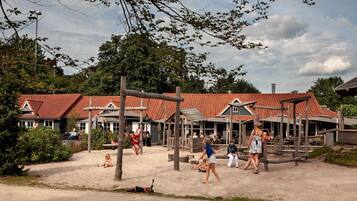 A beachside playground with swings and a sandbox, surrounded by buildings with red-tiled roofs.