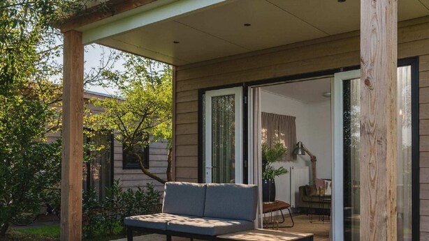 Een modern buitenzitgedeelte met een bank en een tafel, een houten pergola en een glazen schuifdeur naar een binnenruimte.
