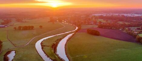 Eine Vogelperspektive auf eine ländliche Landschaft bei Sonnenuntergang, mit Feldern, einem gewundenen Kanal und Gebäuden in der Ferne.