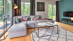 A modern living room with a grey sectional sofa, a coffee table, and a TV on a stand. There are large windows with curtains, a painting on the wall, and a view of greenery outside.