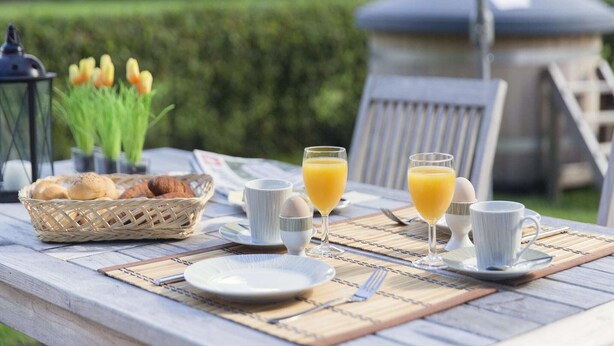 Une table en bois dressée à l’extérieur pour le petit-déjeuner, avec un panier en osier rempli de pain, deux verres de jus d’orange et une tasse de café.