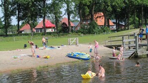 Une scène au bord du lac, avec des gens qui se baignent et jouent sur la plage, un quai et des maisons en arrière-plan.
