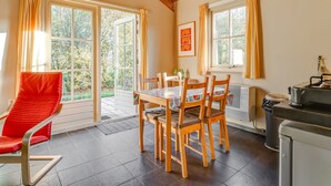 A cozy dining area with a table set for four, surrounded by wooden chairs. Large windows with curtains allow natural light and views of greenery outside. - Holiday home with terrace (Lage Zwaluwe)