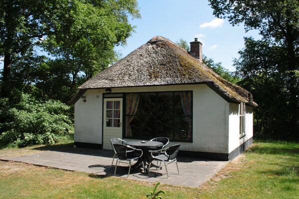 A small thatched-roof cottage with a door and windows, a paved patio, and a table with chairs.