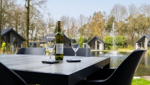 A table with a bottle of wine and two glasses, set outdoors with a view of houses and a pond.
