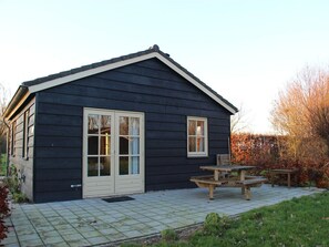 Una casa de madera oscura con una puerta principal, dos ventanas y un patio con una mesa de picnic y bancos.