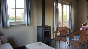 A cozy room with a fireplace, wicker chairs, a white table, and large windows with curtains. - Cozy vacation home with terrace (Lage Zwaluwe)