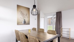 Een eetruimte met een houten tafel, zes stoelen, een hanglamp en een schilderij van een vaas met takken.
