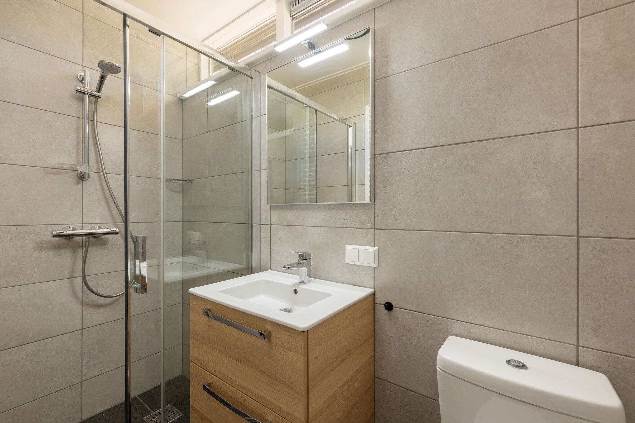 Un bany modern amb plat de dutxa de vidre, un moble de tocador de fusta amb un lavabo blanc i un vàter.
