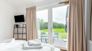 Ein Hotelzimmer mit Bett, Fernseher, einer kleinen Ablage mit Pflanze und Blick auf eine Außenpatio mit Stühlen und Grünpflanzen.
