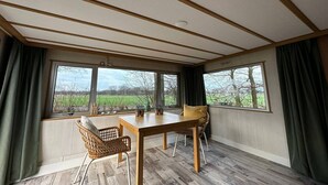 Une pièce avec une table et des chaises en bois, de grandes fenêtres avec des rideaux et une vue sur un champ verdoyant.