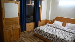 A bedroom with a wooden wardrobe, a bed with floral bedding, and a window with blue curtains. - Taj Guest House (Manali)