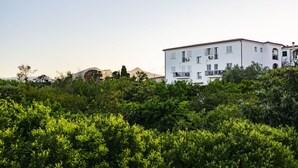Un edificio blanco con varios balcones y unidades de aire acondicionado, rodeado de abundante vegetación.