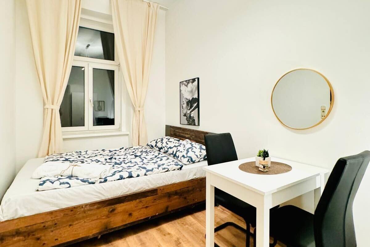 A small, well-lit room with a bed, a table, and a chair.