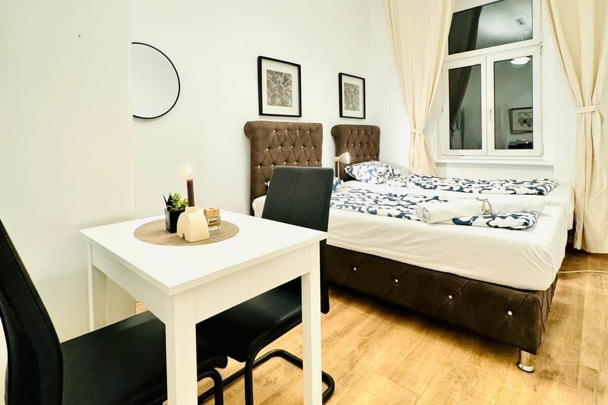 A compact bedroom with a single bed, a small table with a chair, a bedside lamp, and a window with curtains.