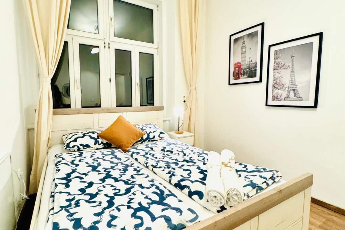A neatly made bed with a patterned bedspread, a single brown pillow, and a set of towels on the side. There is a window with curtains, a bedside table with a lamp, and two framed pictures on the wall.